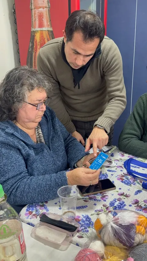 Una mujer mayor acerca la credencial +Cerca a su teléfono, acompañada por un hombre que la guía.
