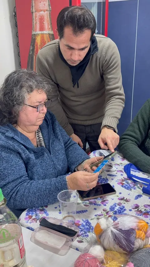 Una mujer mayor prueba la credencial +Cerca con su teléfono mientras un hombre le explica cómo usarla.