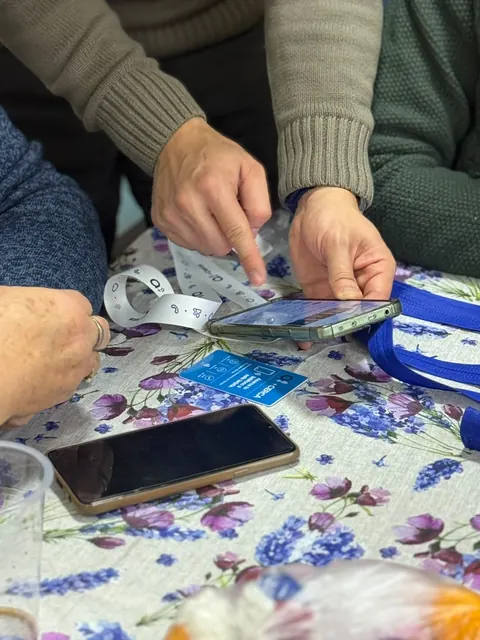 Manos acercan un teléfono a una credencial +Cerca sobre una mesa con mantel floral.