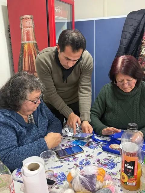Dos mujeres mayores y un hombre revisan juntos sus teléfonos y credenciales +Cerca en una mesa.