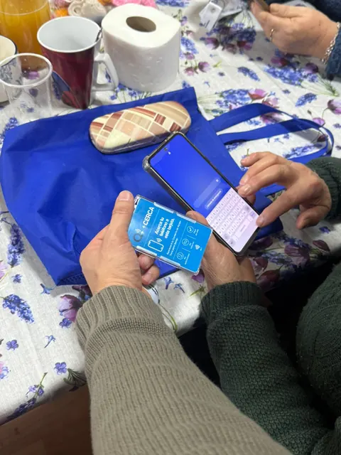 Manos sostienen un teléfono y una credencial +Cerca sobre una mesa con una bolsa azul.