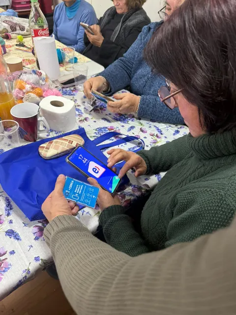 Una mujer mayor acerca un teléfono a la credencial +Cerca, con otras participantes al fondo.