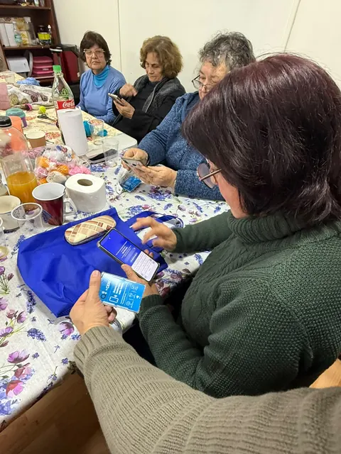 Participantes sentadas en una mesa con teléfonos y credenciales +Cerca; otras personas al fondo.