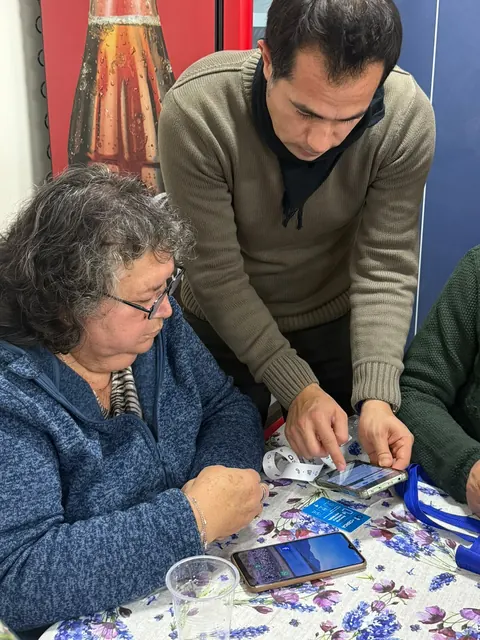 Una mujer mayor observa su teléfono y la credencial +Cerca mientras un hombre le indica los pasos.
